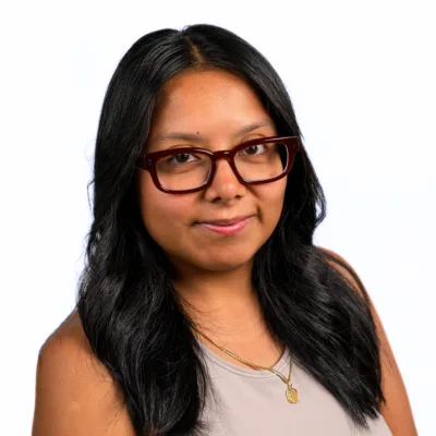 Portrait of a woman with long black hair wearing burgundy glasses and a beige sleeveless top, facing slightly left.